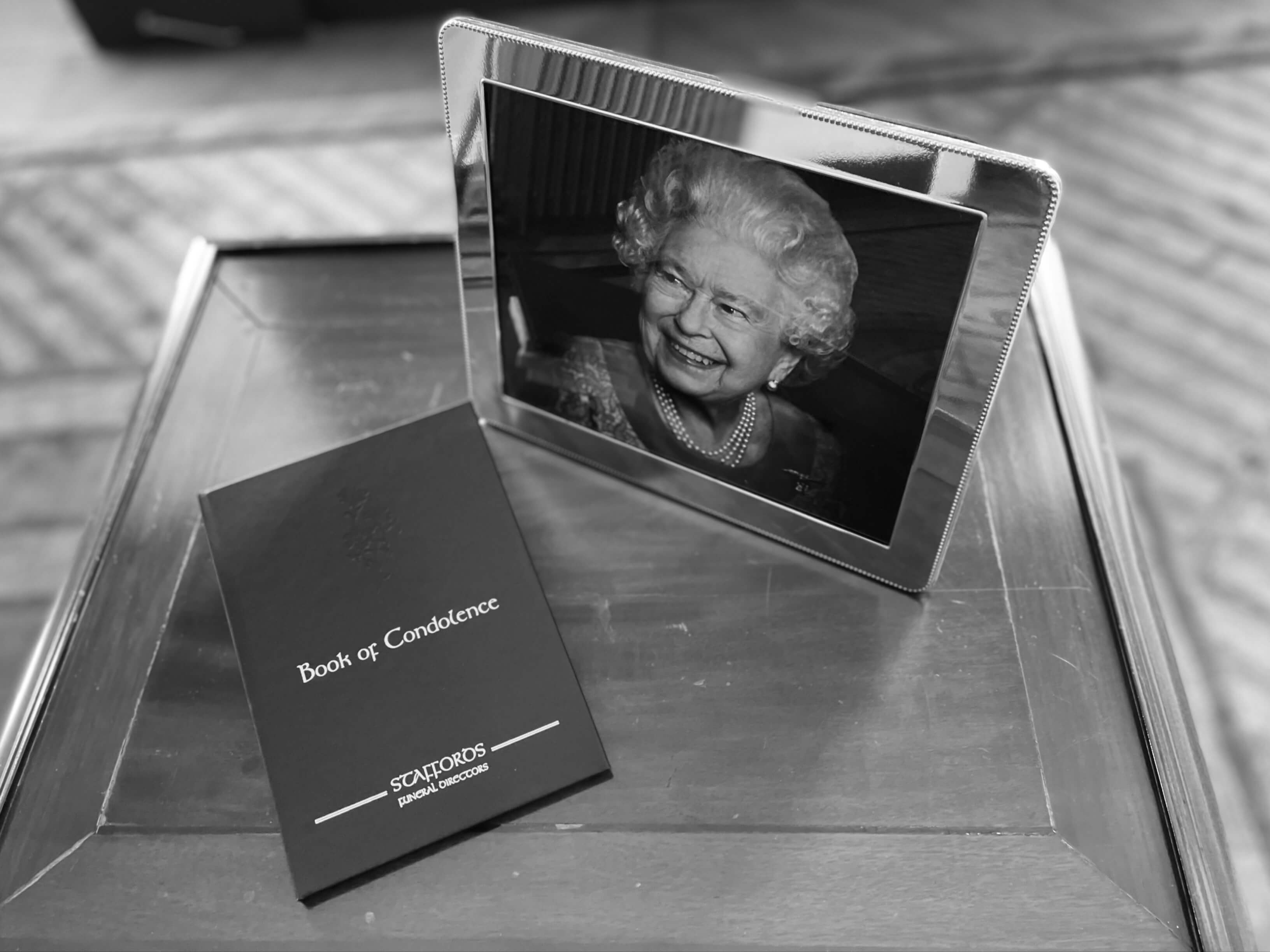 Black and white photograph of a framed head and shoulders photograph of Queen Elizabeth II in a silver frame, accompanied on a table by a black-covered book of condolence