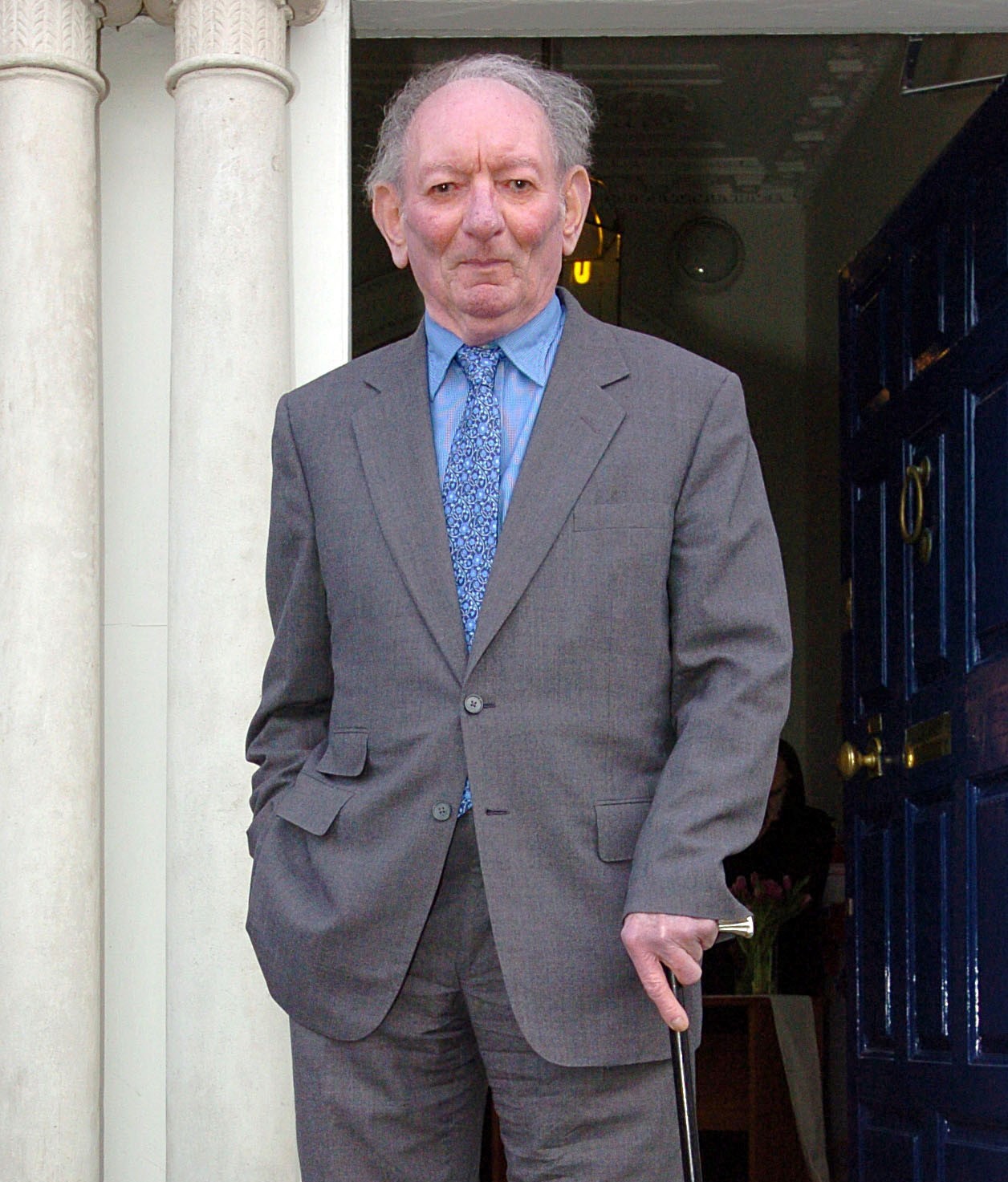 Colour medium distance photograph of Brian Friel, a former Member of Seanad Éireann, in a suit and leaning on a walking stick. He is photographed outside a doorway.