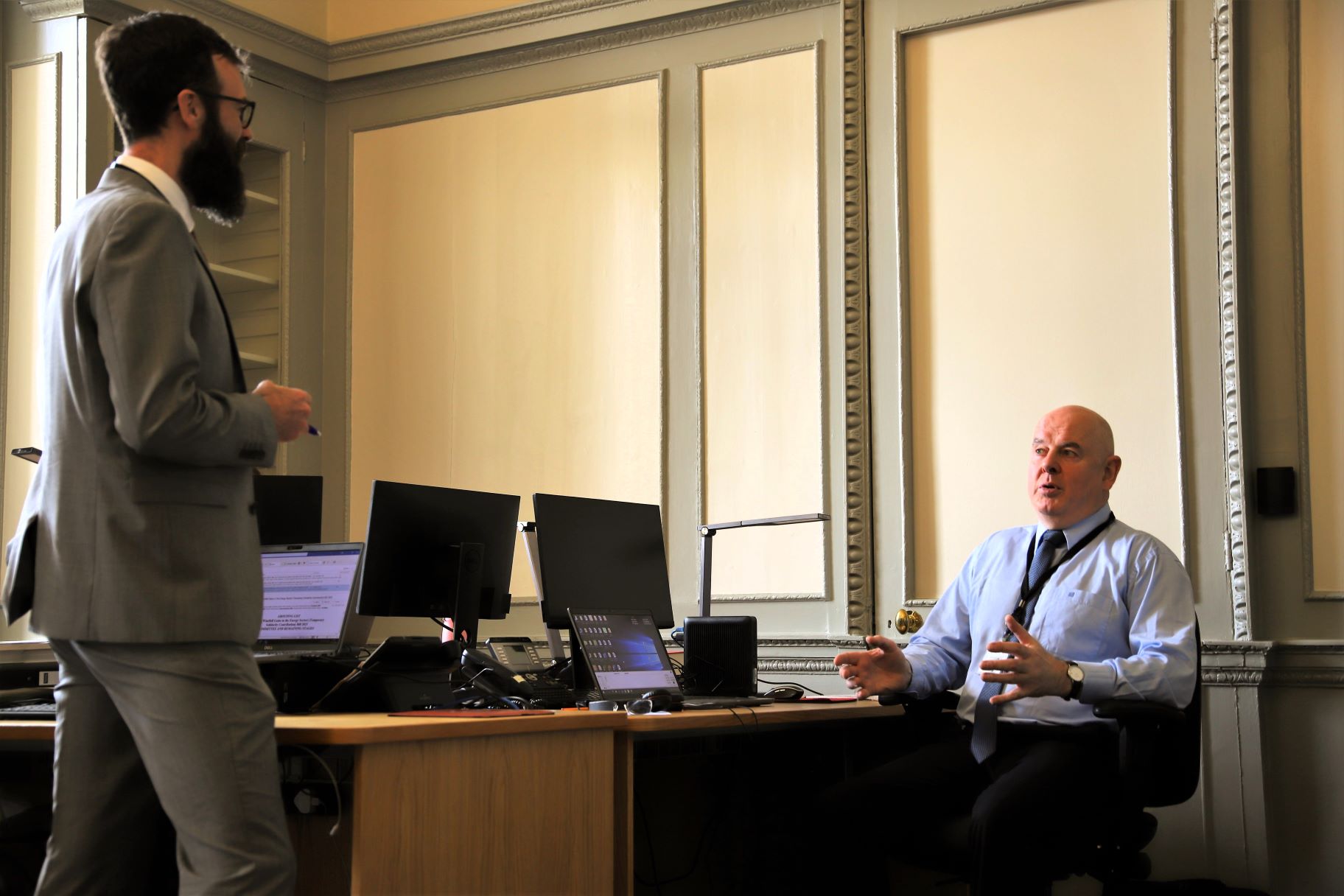 A parliamentary reporter is standing in his office speaking to a colleague, who is seated
