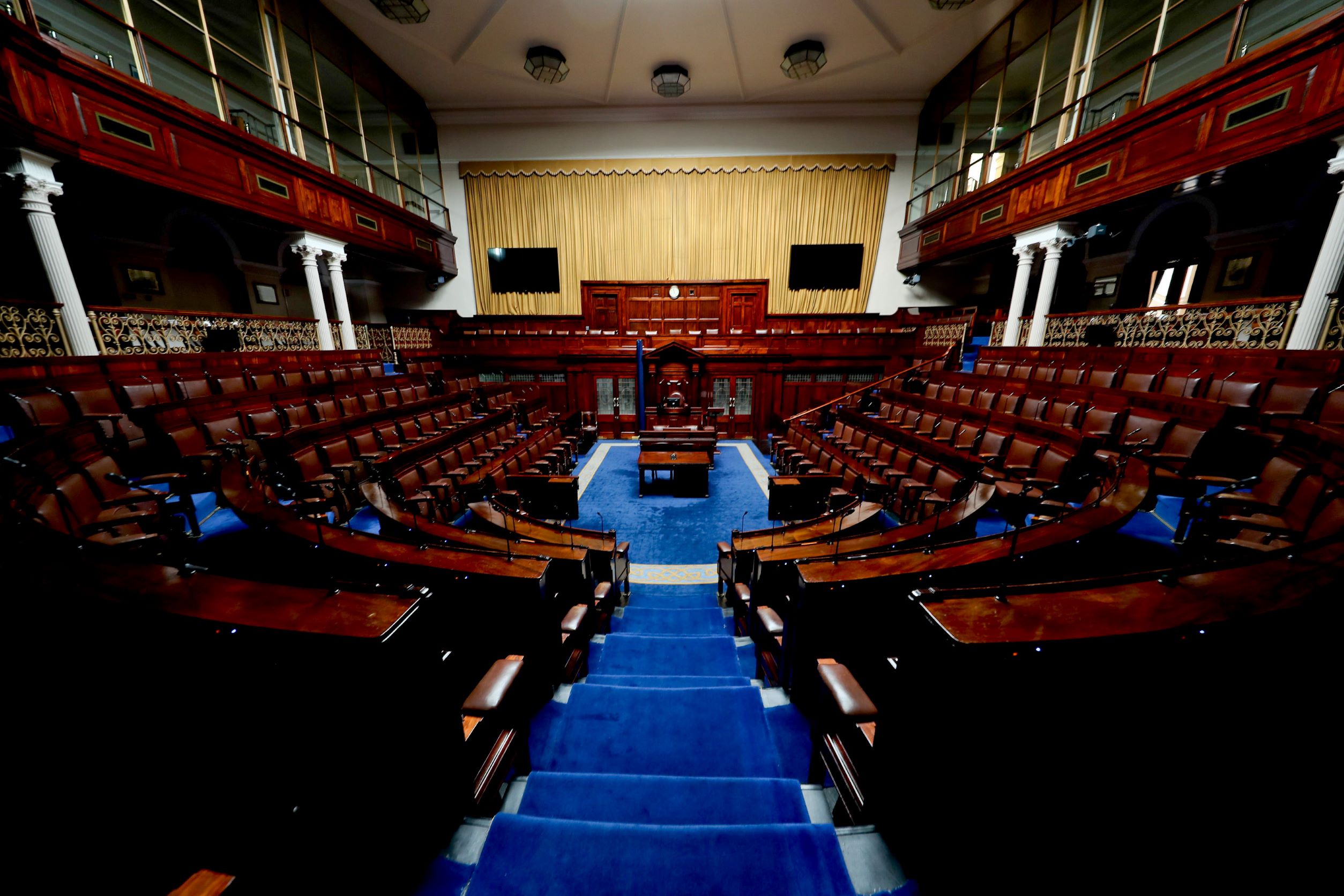 A colour photograph of an empty Dáil Chamber, looking directly towards the Ceann Comhairle's seat from the top of steps