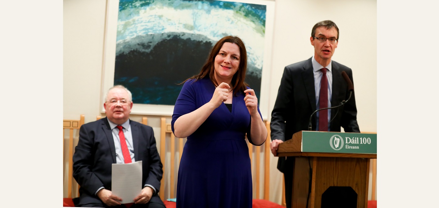 Clerk of the Dáil speaking at a podium with an ISL interpreter providing sign language